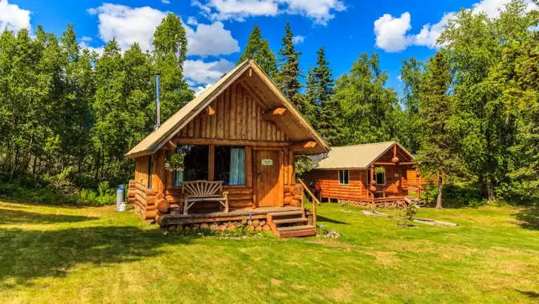 Exterior of guest cabins at Winterlake Lodge with log cabin construction, front porch with a bench & grass & forest surrounding.