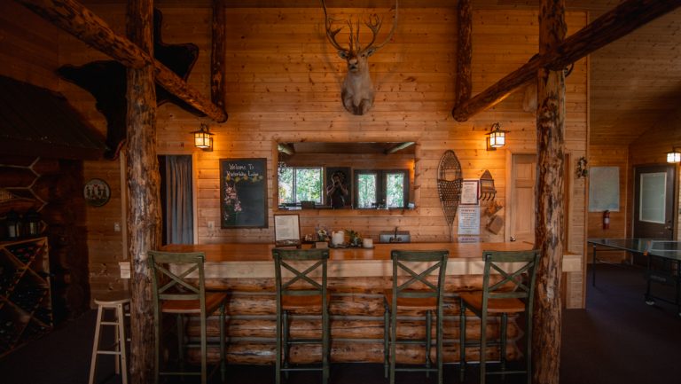 Empty Winterlake Lodge main lodge bar with taxidermized deer, wood posts & beams, rustic barstools & low lighting.