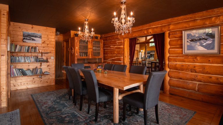 Winterlake Lodge dining room with long wood table, dark upholstered chairs, crystal chandeliers, bookshelf & log cabin construction.