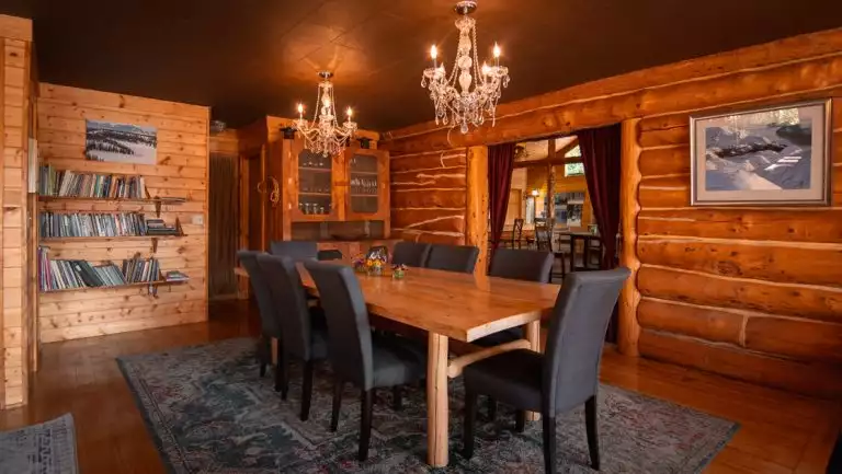 Winterlake Lodge dining room with long wood table, dark upholstered chairs, crystal chandeliers, bookshelf & log cabin construction.