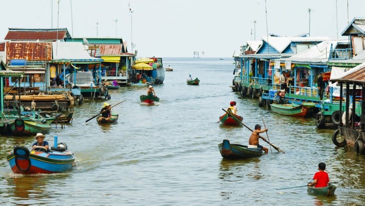 Two rows of people in canoes paddle down the Mekong River through a village in Cambodia