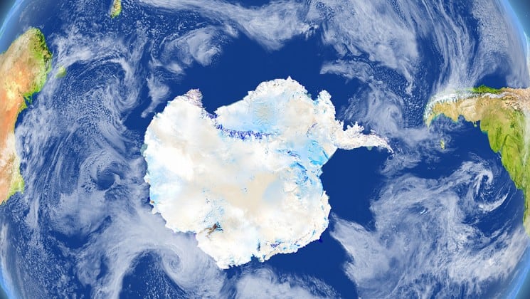 A satellite image of the white continent, antarctica, surrounded by blue ocean and green land with clouds swirling all around