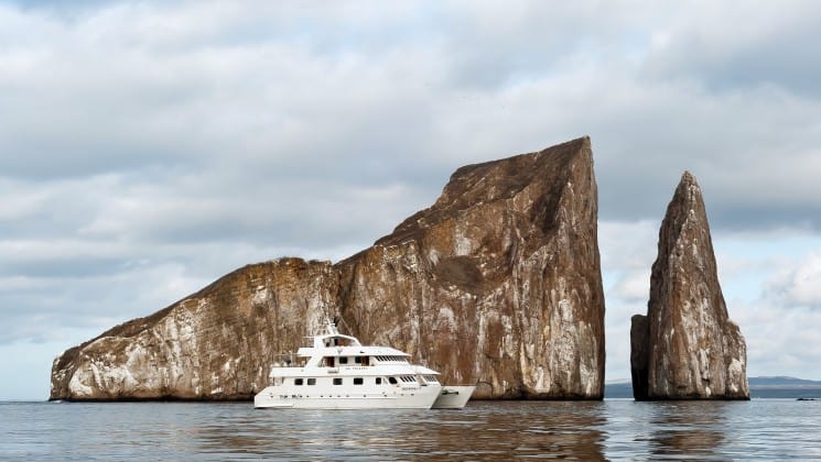 The seaman journey cruise, a luxury yacht, motors across a calm ocean in front of rocks jutting out of the water at the Galapagos islands