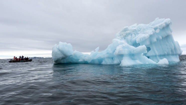 An iceberg rises out of a grey ocean next to an inflatable boat full of passengers from the expedition around spitsbergen