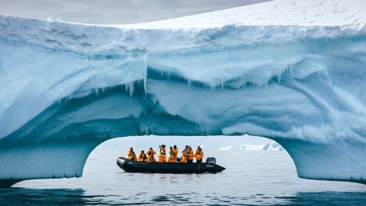 A zodiac boat is visible beneath an iceberg that's arching over the ocean, in antarctica