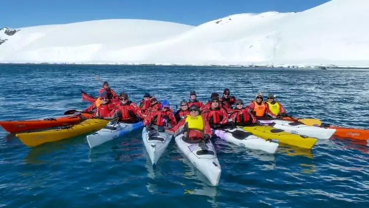A group of kayaks pull up together in the antarctic peninsula with an iceberg in the background