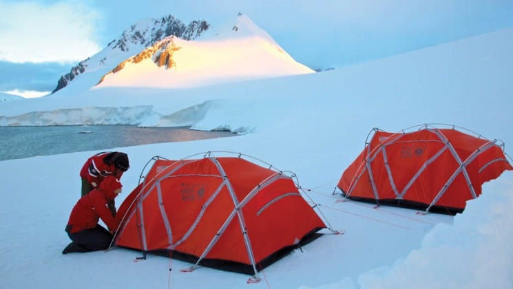 A person stands outside two tents set up on a snowy field in antarctica