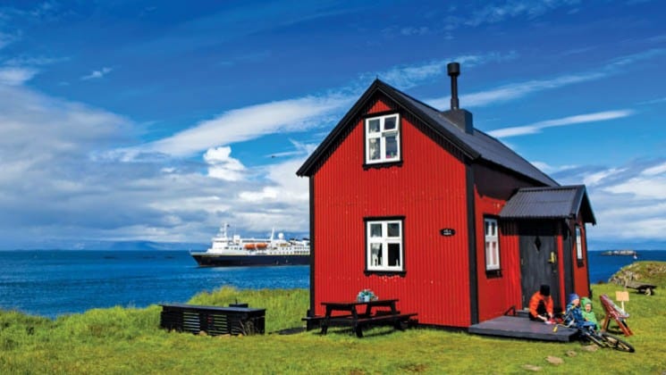 a bright red house on a green pasture in iceland while the national geographic explorer ship motors on the sea in the background