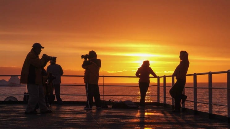 Passengers from the expedition cruise classic antarctica look over the railing of the ship while the sun sets in the background