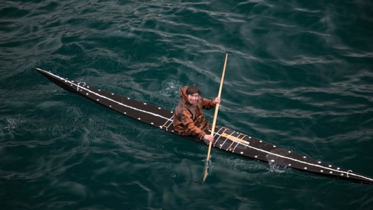a man wearing a brown jacket paddles a traditional wooden kayak in greenland