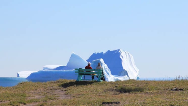 a person sits on a bench on the tundra in the arctic circle, looking at an iceberg in the distance