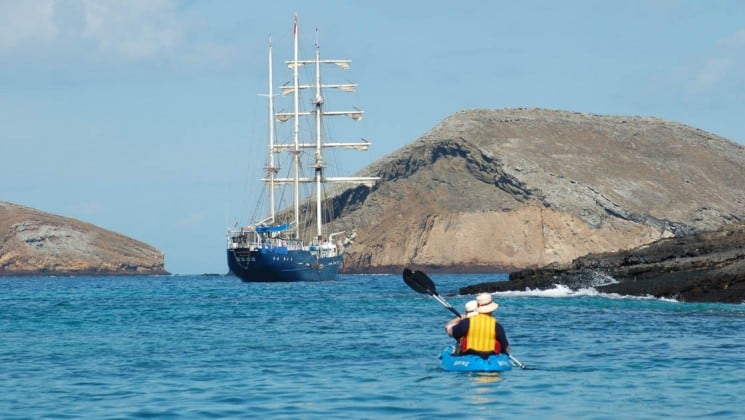 A couple in yellow lifejackets and white hats kayaks across the ocean toward the S/S Mary Anne, a sailing ship anchored next to the Galapagos Islands