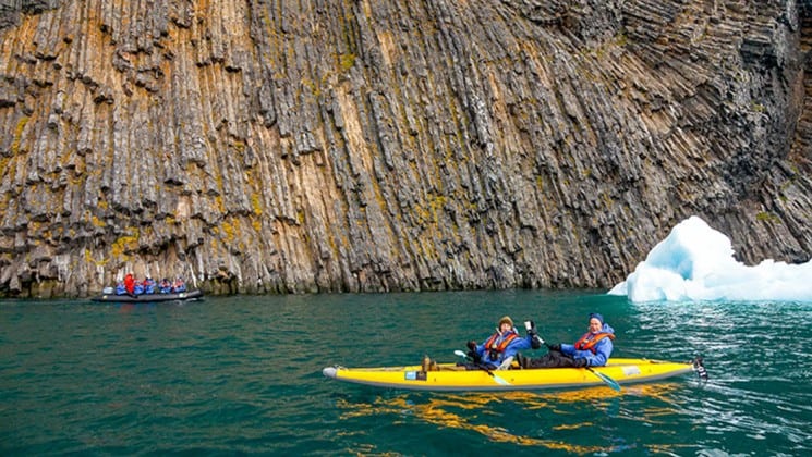 a kayak paddles through greenish-blue water next to a cliffside on the national geographic voyage to the northeast passage