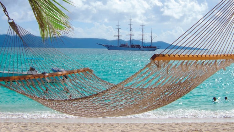 an empty hammock hangs on a white sand beach with the tropical caribbean ocean and the sea cloud small ship sailing in the background