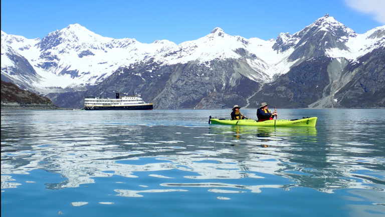 two travelers kayaking inside Glacier Bay National park, the SS Legacy small ship folats behind them and in the background a snowy mountain range