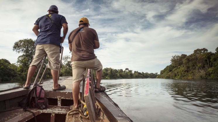 Two men standing at the bow of a small boat looking ahead as they go down a river in the Peruvian Amazon
