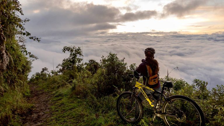 Bike riding activity on an Ecuador land tour. A female guest wearing a helmet stands next to her yellow bicycle at the very top of a lush green trail, lookout out over the endless sea of white clouds