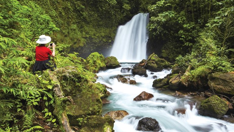 A female traveler in a red shirt and white wide brimmed hat kneels in lush green forest photographing a waterfall in the jungle of Costa Rica.