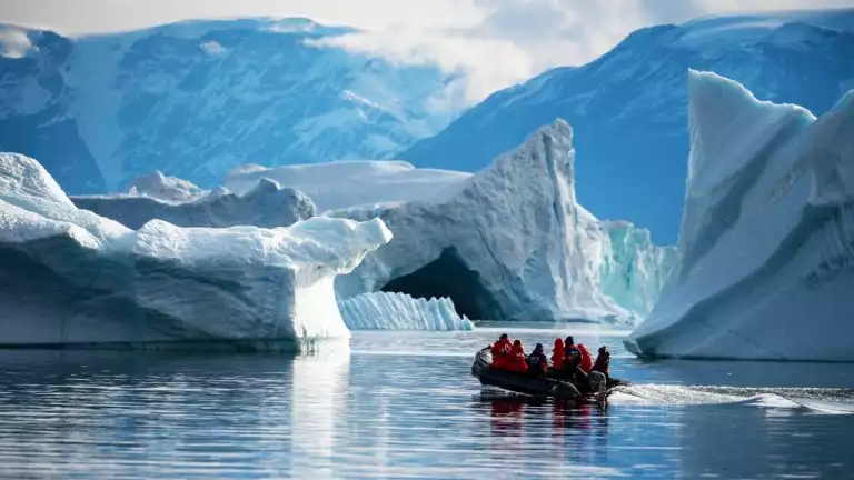 A zodiac navigates large icebergs in calm still waters with snowy mountains off in the distance
