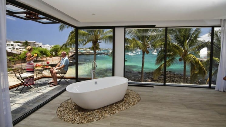 standalone bathtub on wood floor of a luxury galapagos hotel with large windows behind it overlooking the ocean and travelers sitting outside