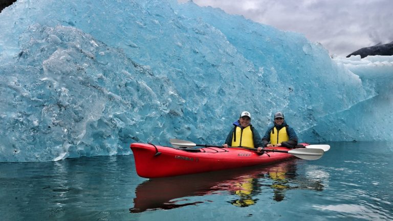 Two women wearing yellow life jackets pose with their paddles in a red double kayak in front of an teal blue iceberg in Alaska.