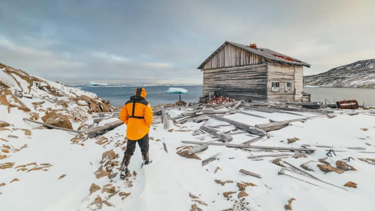 Polar traveler in yellow jacket walks over snowy ground toward an old wooden hut during the Jewels of the Russian Arctic small ship cruise.