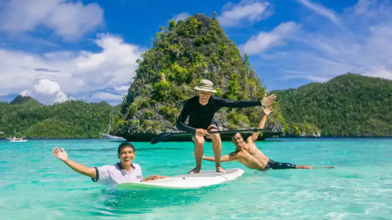 three travelers playing on a stand up paddleboard in crystalline water with vibrant green islands behind them on a sunny day in indonesia