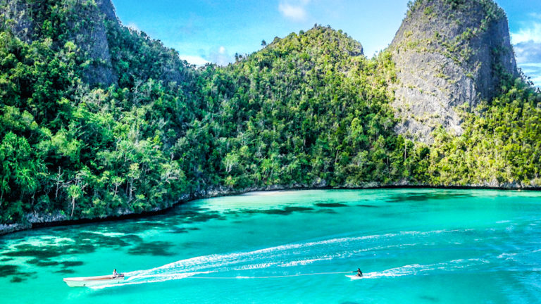 gorgeous turquoise cove surrounded by jungle in indonesia with a small ship pulling a traveler on a stand up paddleboard