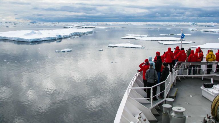 A group of people stand on board the ship for the national geographic white continent voyage to look at icebergs in the ocean in antarctica