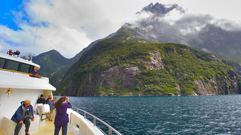Several expedition cruise guests on the bow of their ship in New Zealand photographing the tall green coastal mountains with their peaks in the clouds.