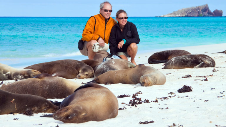 a man and a woman crouch low to pose for a photograph with the sea lions asleep on a white sand beach with turquoise water in the background at the Galapagos islands