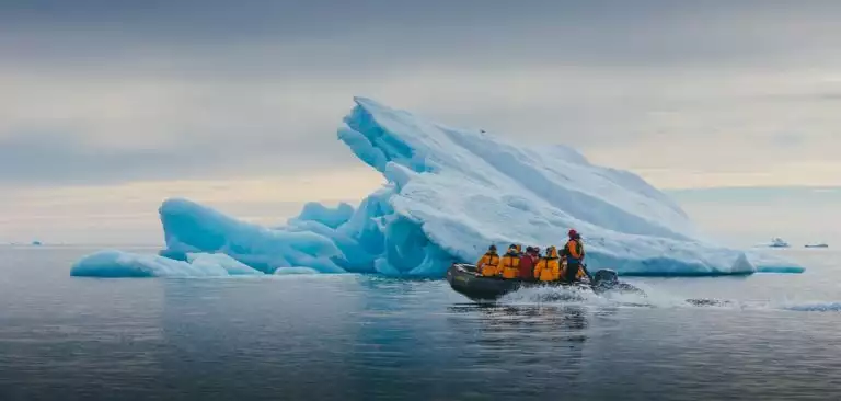 Small group of expedition ship guests wearing orange parkas on a zodiac excursion with their guide getting close to an impressive iceberg in the Arctic.
