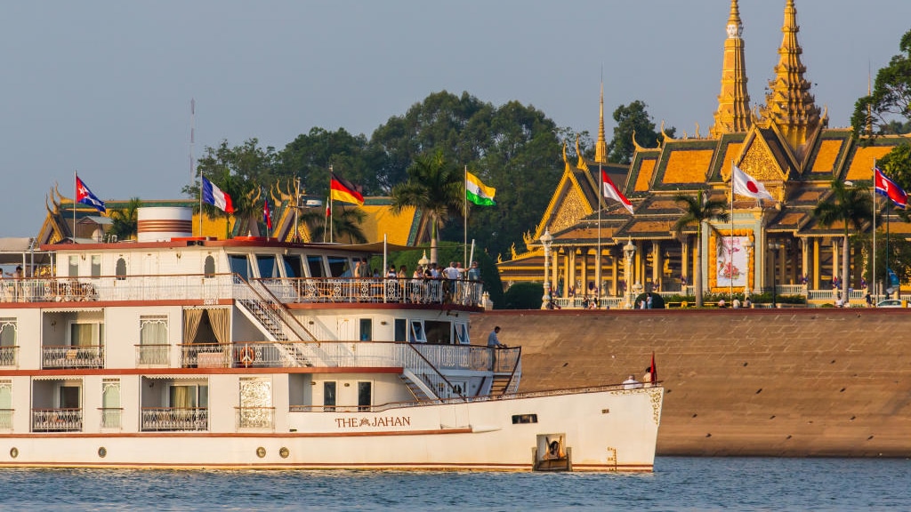 The Jahan river boat cruises the Mekong river in Cambodia with guests on the bow, an intricate golden temple is seen onshore.