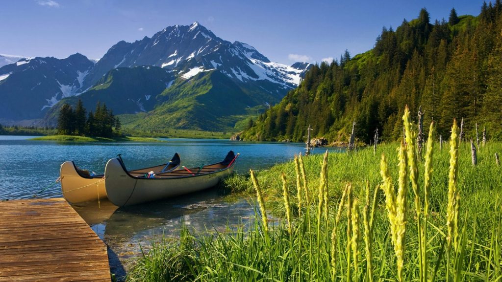 In Alaska. two canoes are tied up to a dock that extends into a blue lake surrounded by jagged snow capped mountains and dense forested hillside.