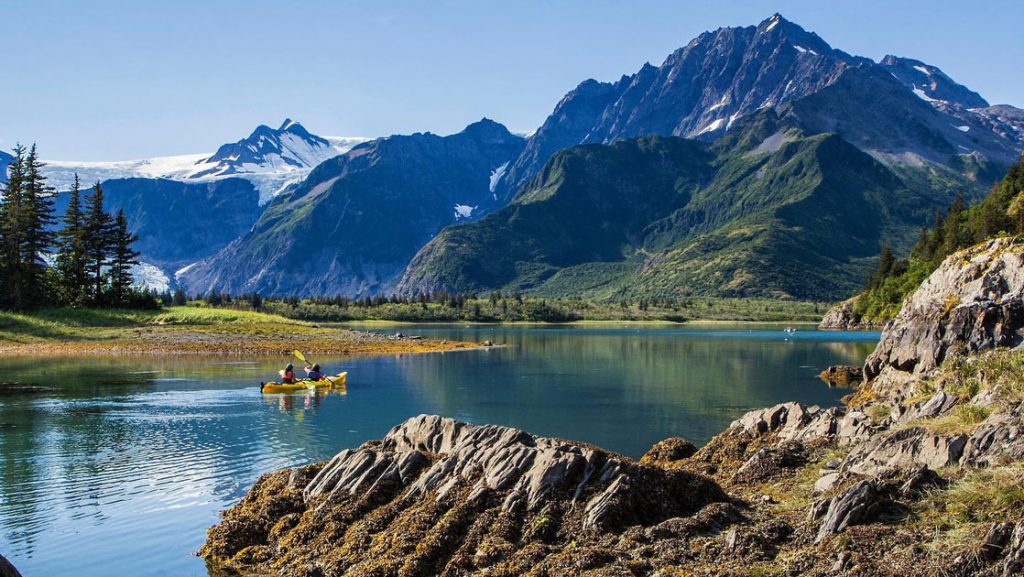 Tandem kayakers paddle glassy waters at Pedersen Lagoon, beside rocky shoreline & craggy green mountains on the Alaska Grand Adventure.