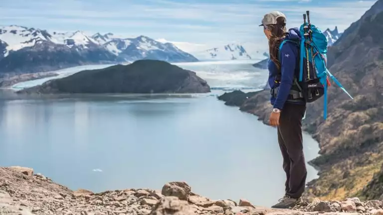 Young woman wearing a day pack with trekking poles stands on a rocky summit looking down on a lake on the Torres del Paine Trek.