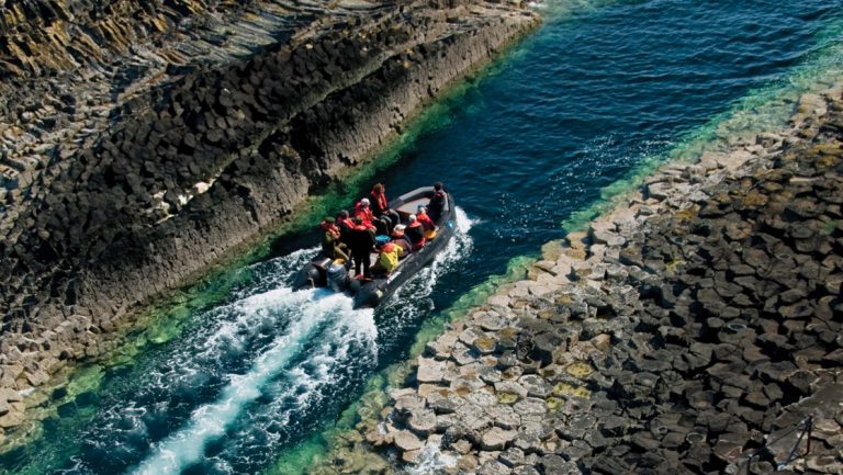 Zodiac boat with Scotland travelers cruises in narrow blue-green water between 2 shores with angular gray rocks.