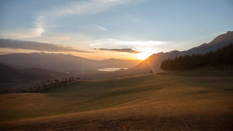 The sun it setting over the mountain range on Ecuador's Andes, rolling hills open field and a lake are glowing.