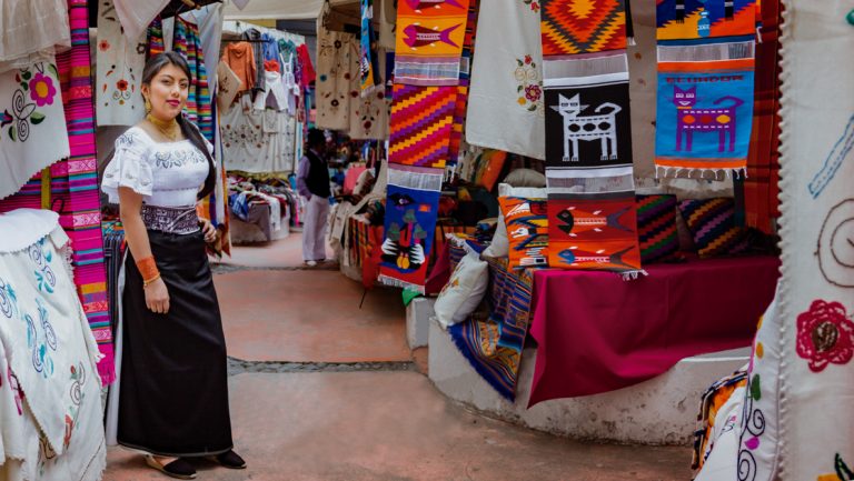 Woman shopkeeper stands in open-air market amongst brightly colored woven textiles, seen on the Andes Highland Haciendas tour.