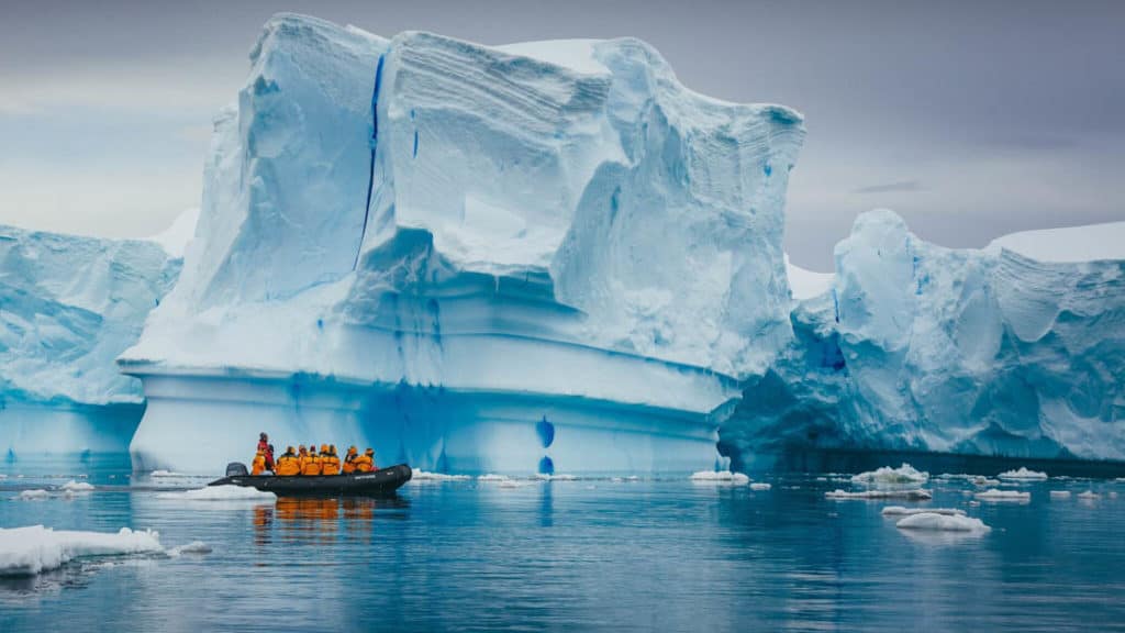 adventure travelers wearing yellow jackets in a zodiac skiff cruise in front of a large iceberg on an overcast day in antarctica