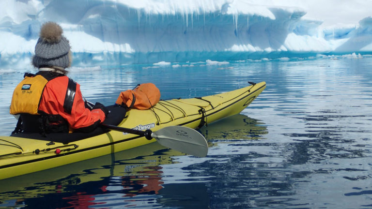 adventure traveler in a yellow kayak sits in calm water looking at an iceberg on the antarctic explorer small ship cruise trip