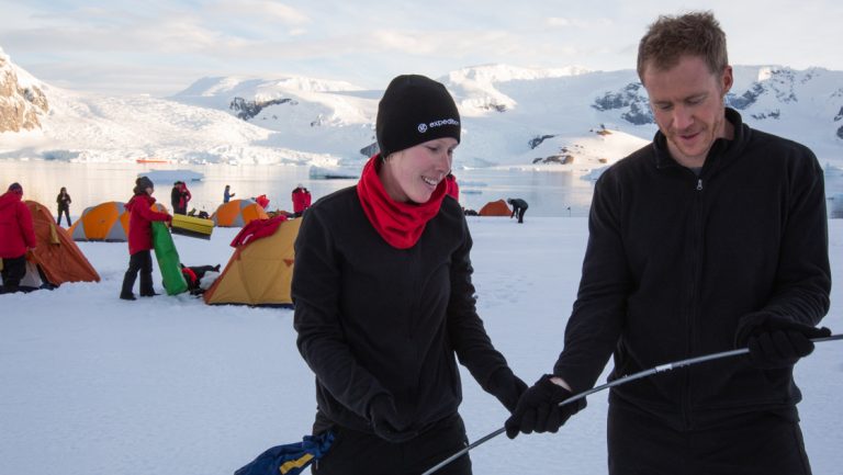 Man & woman in black clothing put together tent poles with others setting up tents in the snow behind them, in Antarctica.