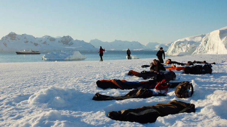 Sleeping bags in bivvy sacks at sunset lay atop Antarctica's snow-covered landscape with a small ship in the background.