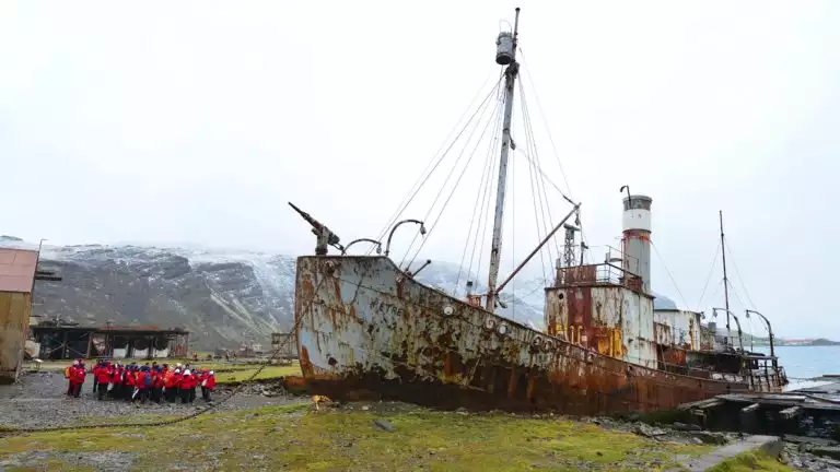 Group of travelers wearing red parks on a land excursion looking up at an abandoned rusty ship in Antarctica.