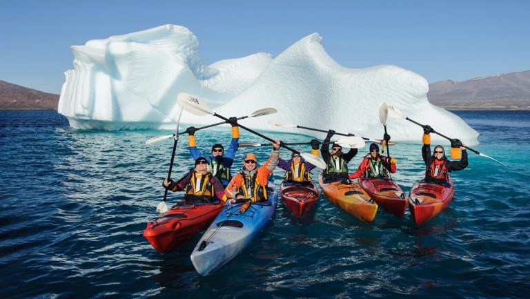 A group of kayakers in red orange and blue kayaks hold their paddles in the air as they float in front of a large iceberg in Antarctica