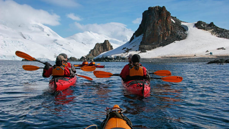 group of adventure travelers kayak on a sunny day in antarctica with large mountains in the distance