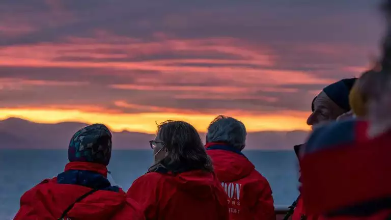 Travelers in red jackets watch the pink and yellow sunset off small cruise ship