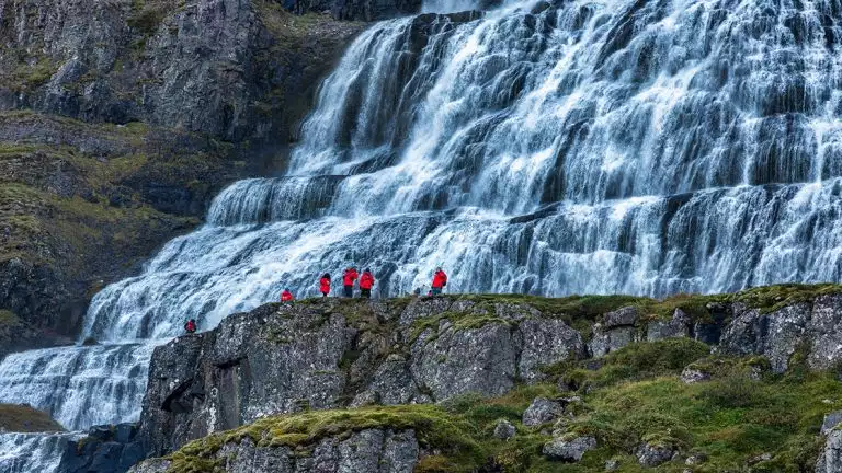 Hikers dressed in red jackets navigate massive water walls that tower over them and fill the whole wall in white water