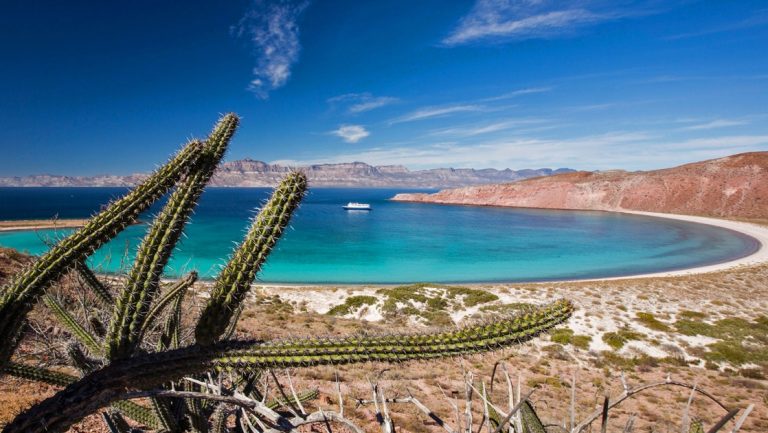 In Baja, a small ship floats inside a bay of teal blue water, partially surrounded by land, a sandy beach shoreline and desert mountain range.