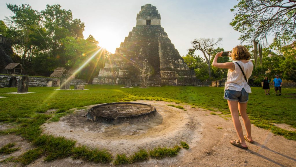 Woman in white top & jean shorts stands below ancient Mayan ruins & takes a photo as sun sets on a Belize & Tikal tour.